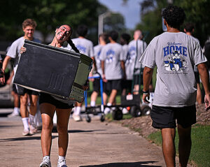 A student smiles while carrying a miniature fridge.