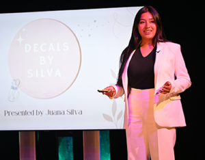 A young Hispanic student with long hair wearing a cream pant suit talks about her presentation.