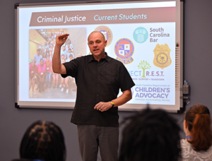 A male criminal justice professor wearing a blue shirt stands in a classroom in front of an digital board.