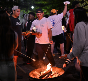 A group of male and female students of different races and ethnicities gather around a fire pit to roast marshmallows.